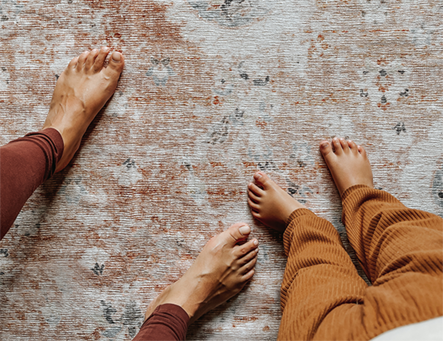 Two pairs of bare feet standing on a patterned rug.